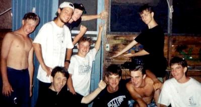 A group of band camp friends outside a cabin; Nick kneels in the center in a white shirt