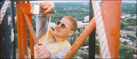 Nick Berg smiling while painting the red steel of a broadcast tower, high above a treeline